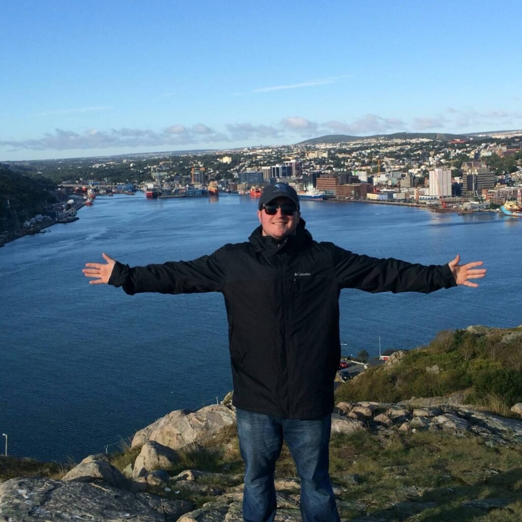 Robby with arms outstretched on a rocky overlook above a harbor and city on a sunny day